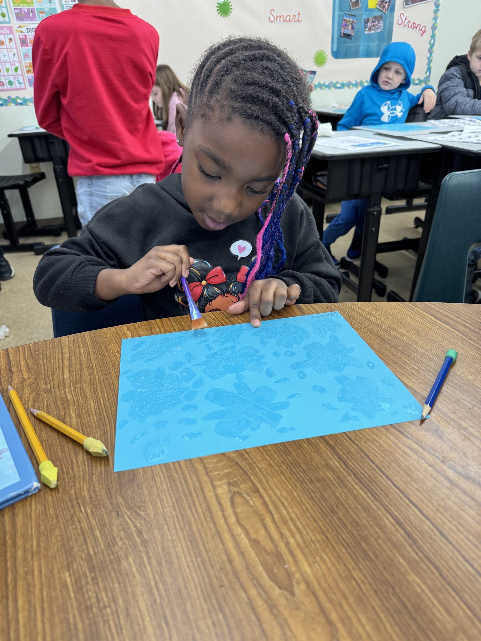 A young student paints on a blue sheet of paper at her classroom table while other students work in the background.