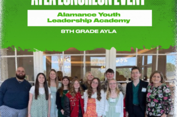 A group of Clover Garden School 8th grade students and staff stand together at the AYLA Luncheon Event for the Alamance Youth Leadership Academy, posing indoors in front of large windows at the Alamance Country Club.