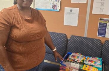 Ms. Kim of Clover Garden AME Church stands beside a box filled with donated school supplies, including notebooks, pencils, crayons, markers, and colored pencils, for Clover Garden School students.