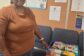 Ms. Kim of Clover Garden AME Church stands beside a box filled with donated school supplies, including notebooks, pencils, crayons, markers, and colored pencils, for Clover Garden School students.
